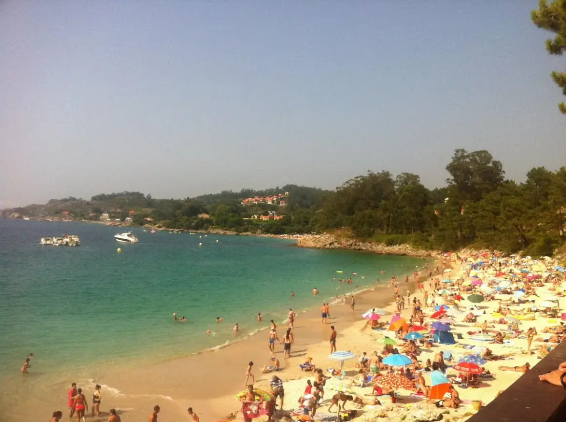 Gente disfrutando del sol y el mar en la playa areacova, con barcos en el agua y colinas verdes al fondo.