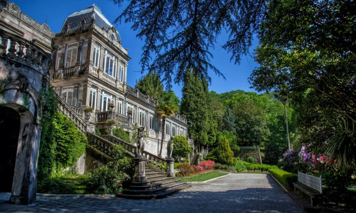 El Pazo de Lourizán, una mansión señorial con escalinatas cubiertas de hiedra y un jardín exuberante, se alza majestuoso bajo un cielo azul.