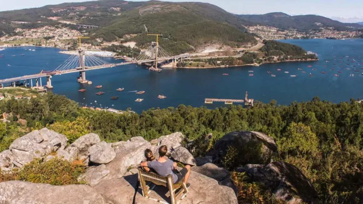 Pareja sentada en un mirador, contemplando el impresionante puente de Rande sobre el mar, con bateas de mejillones y un pueblo al fondo.