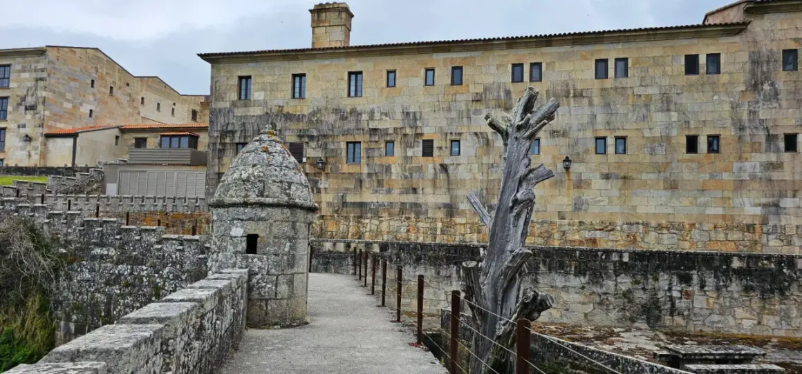 Camino de piedra hacia la fortaleza de Baiona, con una garita y un árbol seco.