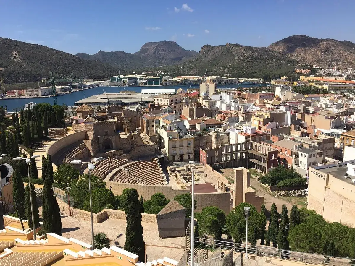 Vista panorámica de Cartagena con el anfiteatro romano y el castillo de la Concepción al fondo, junto a un puerto industrial.