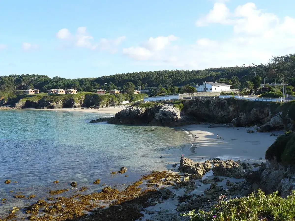 La playa o cantiño Burela se asoma entre rocas y acantilados, con casitas blancas coronando la colina.