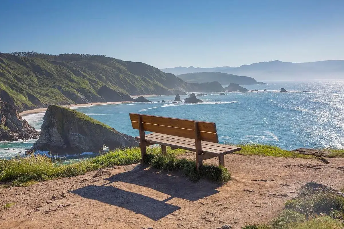 Un banco de madera invita a contemplar el mar azul desde el mirador de Coitelo, con acantilados verdes y rocas emergiendo del agua.