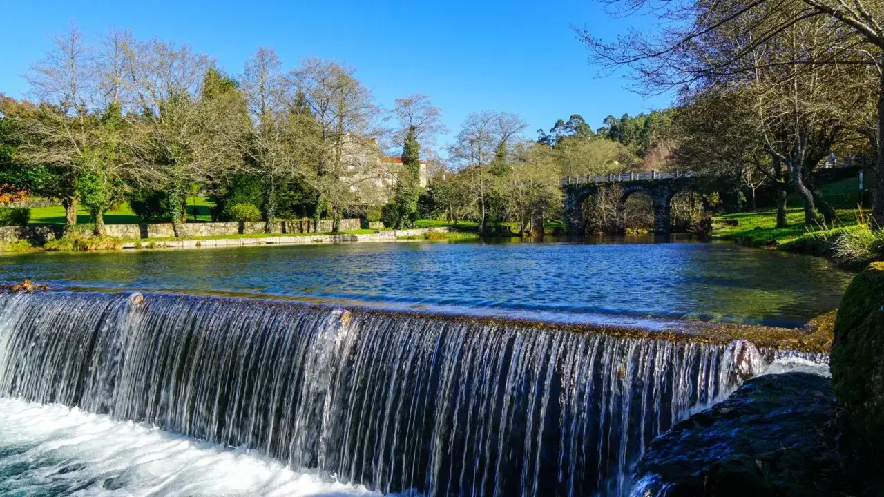 Cascada en la playa fluvial del río Verdugo. Un puente de piedra cruza el río, rodeado de árboles y vegetación exuberante bajo un cielo azul.