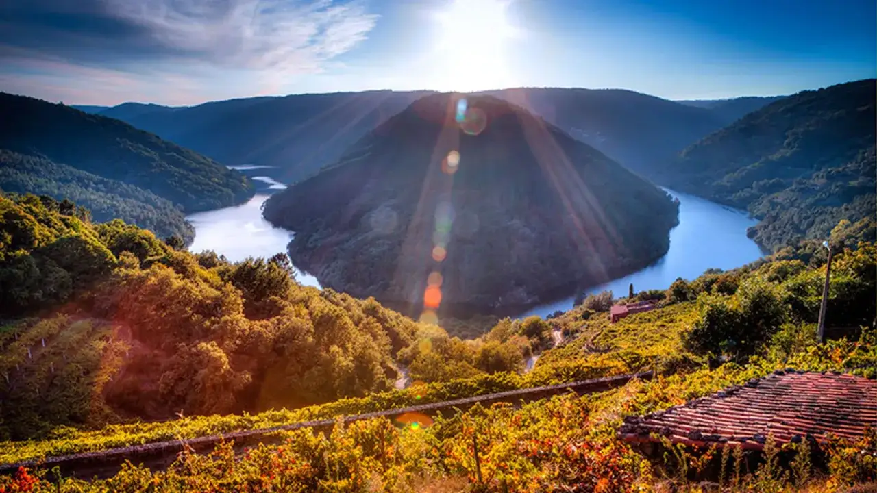 Un río serpentea entre colinas verdes, creando una curva espectacular que parece el cabo do mundo. El sol brilla intensamente, iluminando el paisaje.