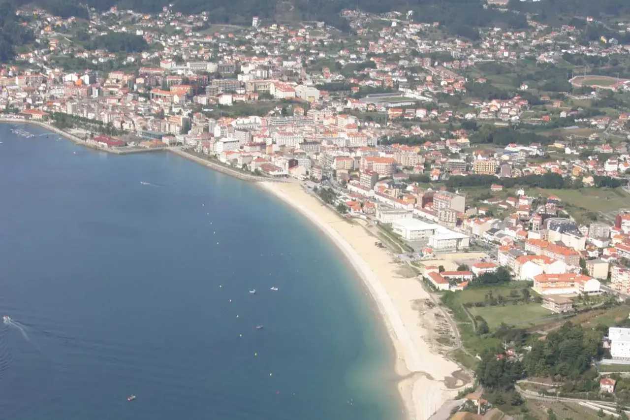 Vista aérea de la playa de Rodeira, con el mar azul, la arena dorada y la ciudad extendiéndose hasta las colinas verdes.