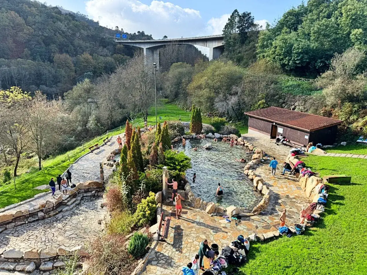 Gente disfrutando de las termas naturales en Ourense, con un puente moderno al fondo.