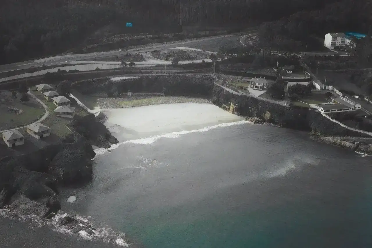 Vista aérea de una playa areoura con casas en la ladera y una carretera al fondo.