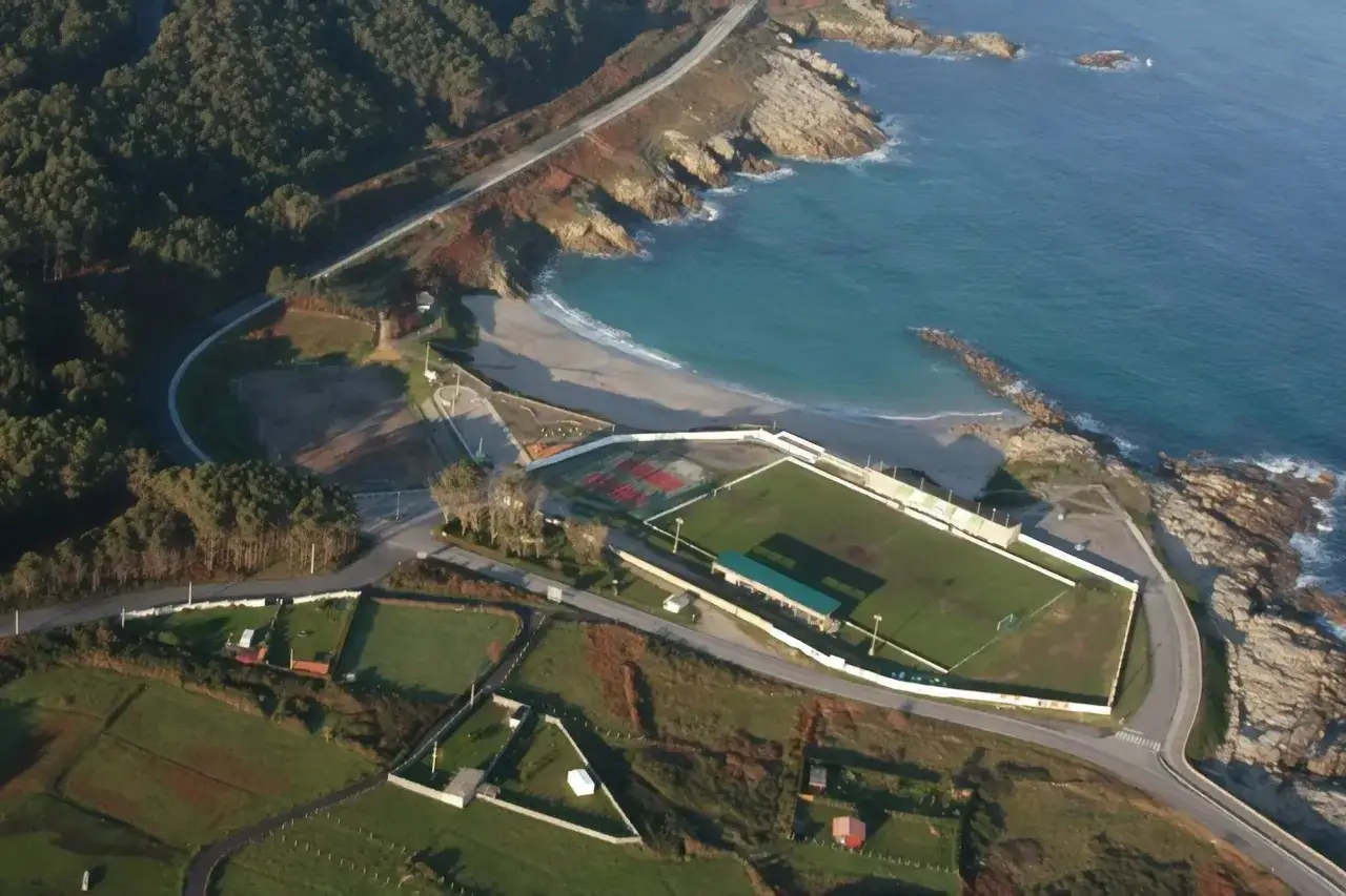 Vista aérea de un campo de fútbol junto a una playa marosa con aguas turquesas y rocas.