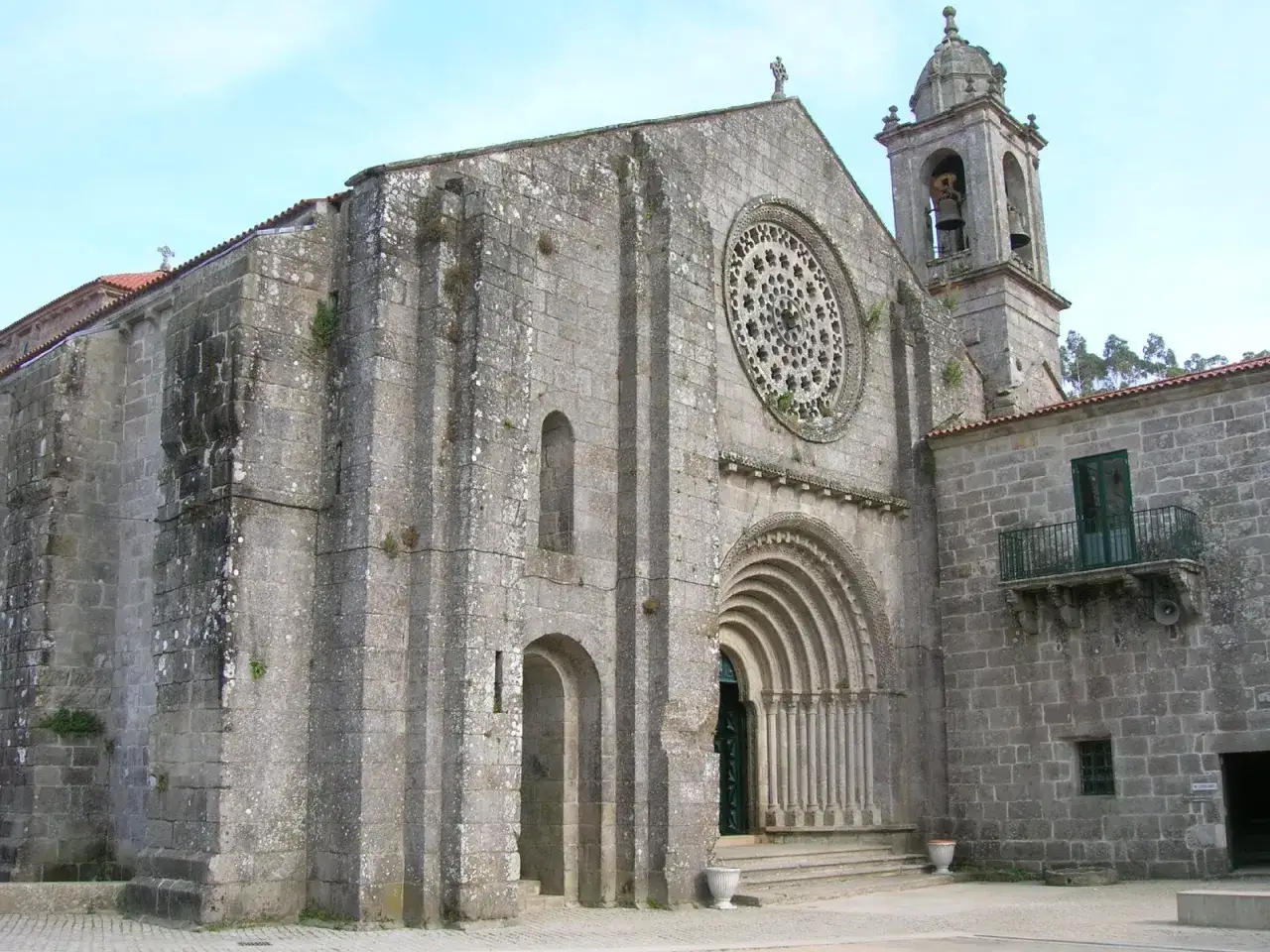 Fachada del monasterio de Armenteira con su rosetón y portada románica. La piedra gris y el cielo azul crean una atmósfera serena.