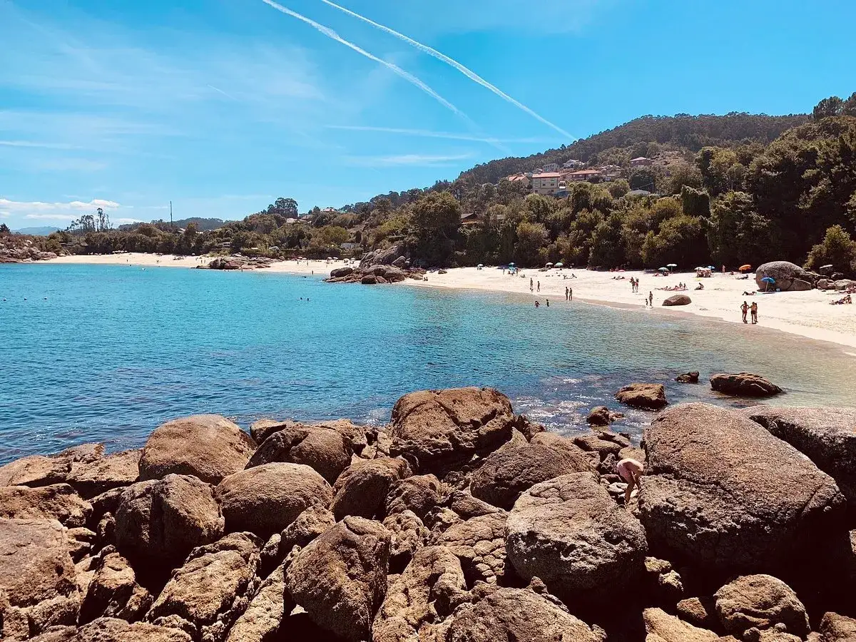 La playa de Castiñeiras luce radiante con arena blanca, aguas turquesas y gente disfrutando del sol. Rocas cubren el primer plano.