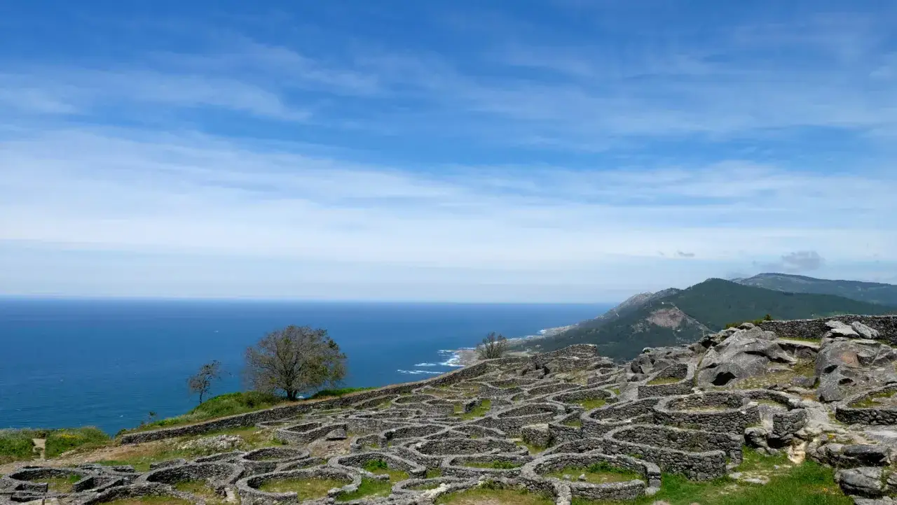 Ruinas circulares de piedra en el monte Santa Tecla, con el mar azul y montañas verdes al fondo bajo un cielo nublado.