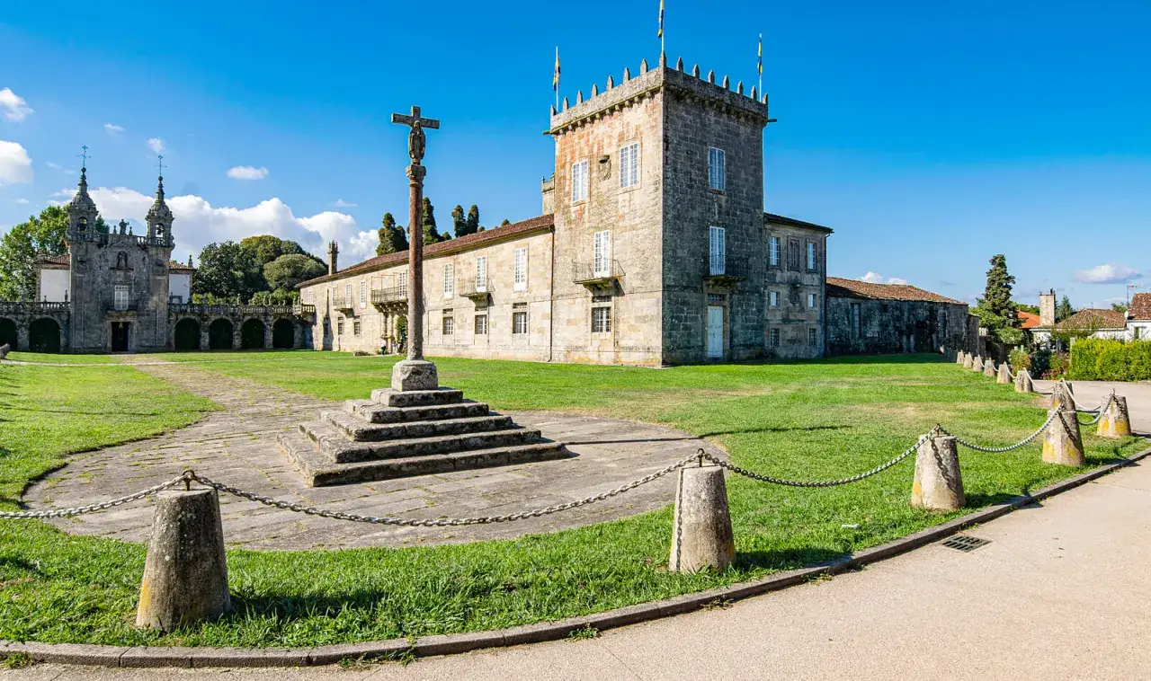 Un cruceiro se alza en el patio de pazos en Galicia, con un edificio histórico y una iglesia barroca al fondo bajo un cielo azul.