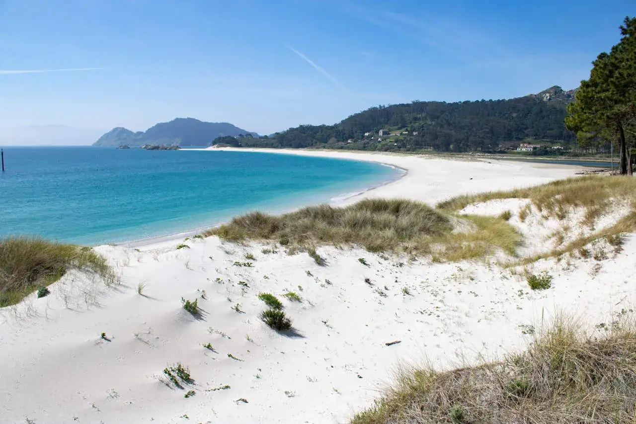 Arena blanca y dunas cubiertas de hierba en la playa de Nerga, con aguas turquesas y colinas verdes al fondo.