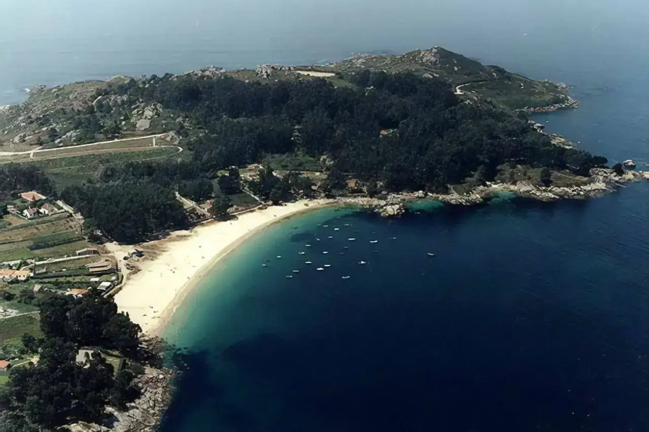 Vista aérea de praia da Mourisca, con arena dorada, aguas turquesas y barcos. Al fondo, colinas verdes y casas.