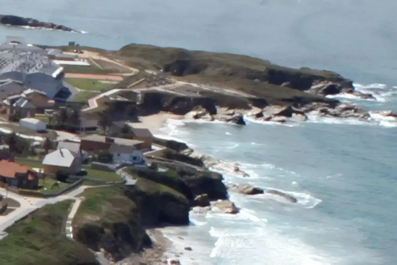 Vista aérea de casas y un complejo deportivo junto a la playa de San Bartolo Barreiros, con el mar agitado y rocas.