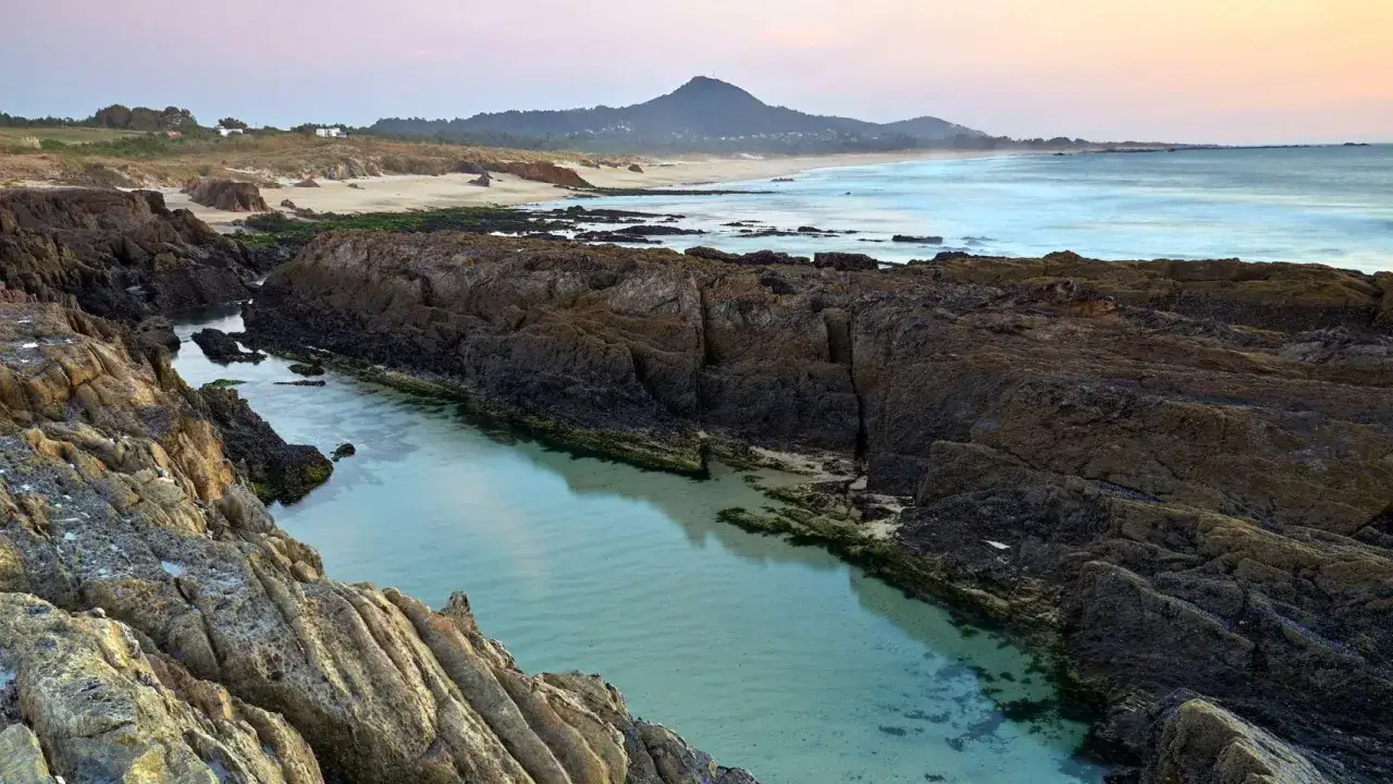 Aguas cristalinas en la playa das Furnas, entre rocas volcánicas y arena dorada, con el mar al fondo y una montaña.