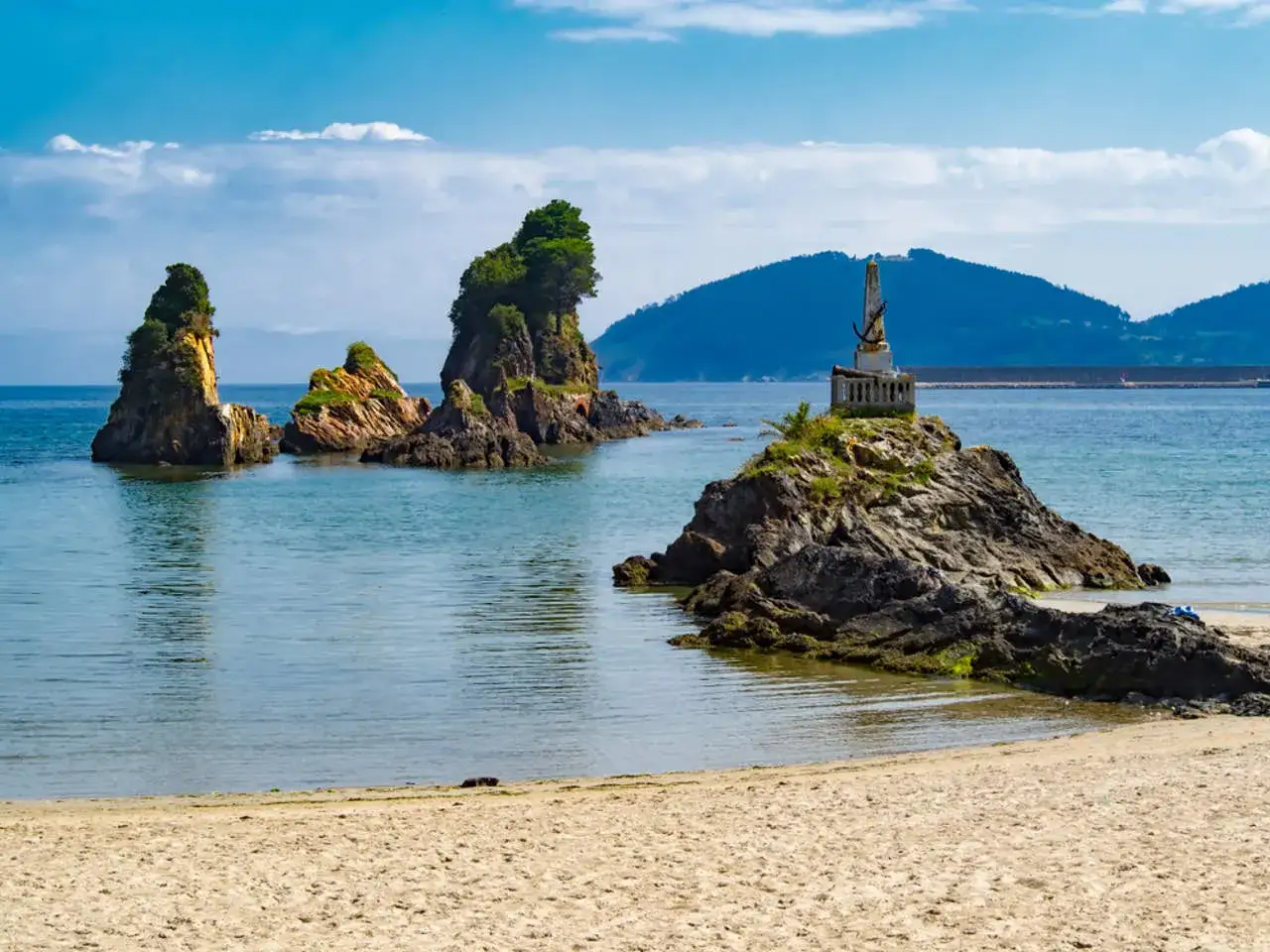 Rocas emergiendo del mar, un monumento en una de ellas, y la arena dorada de las playas cerca de Viveiro bajo un cielo azul.