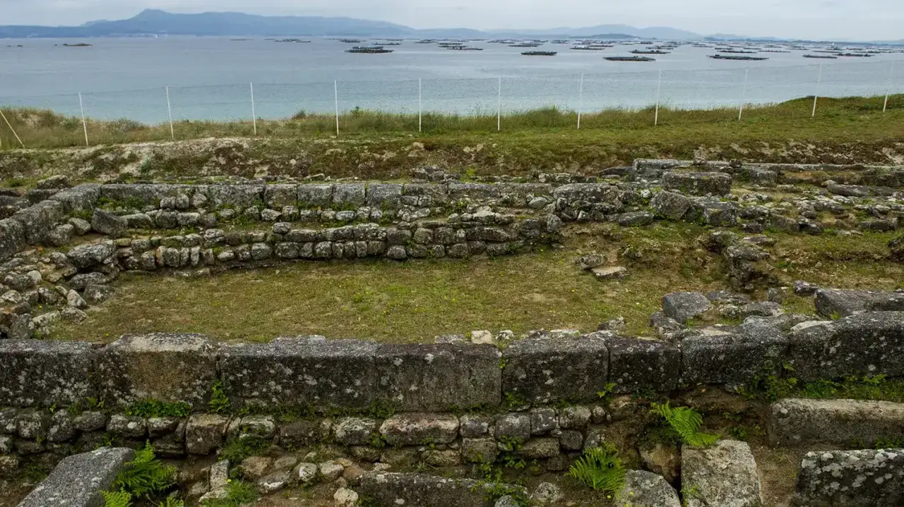 Ruinas de piedra antigua, con el mar y bateas de mejillones al fondo. Un lugar que evoca el adro vello de la historia.