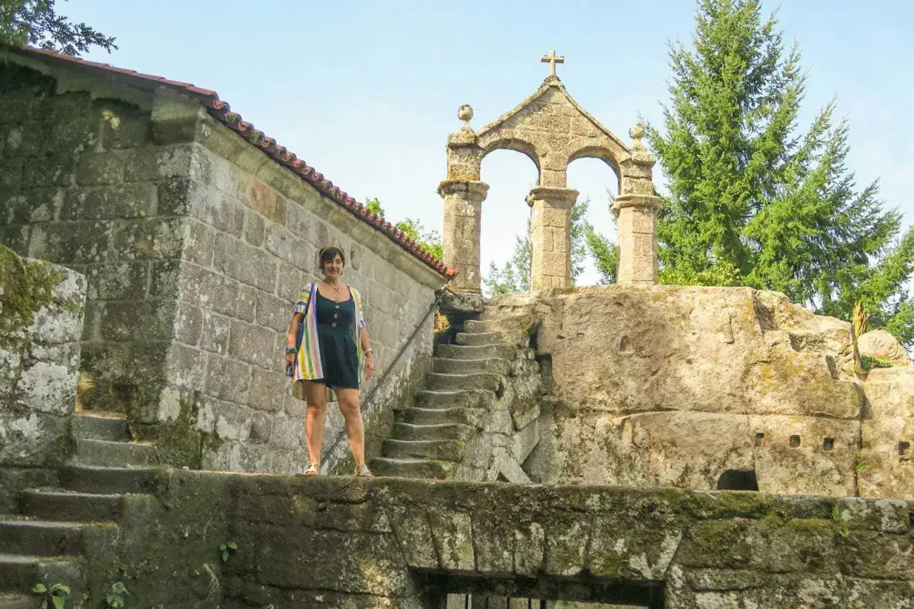 Mujer posa frente a la entrada del monasterio de San Pedro de Rocas, con su arquitectura de piedra y escalinata.