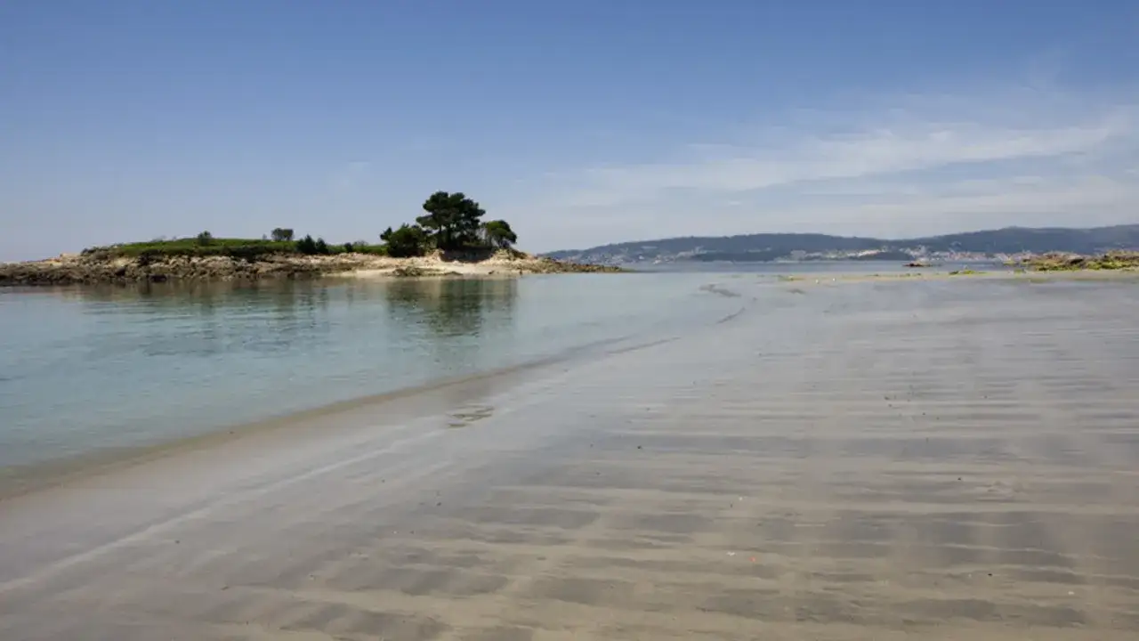 Arena mojada y el mar tranquilo en Praia do Santo, con una pequeña isla rocosa y árboles al fondo.
