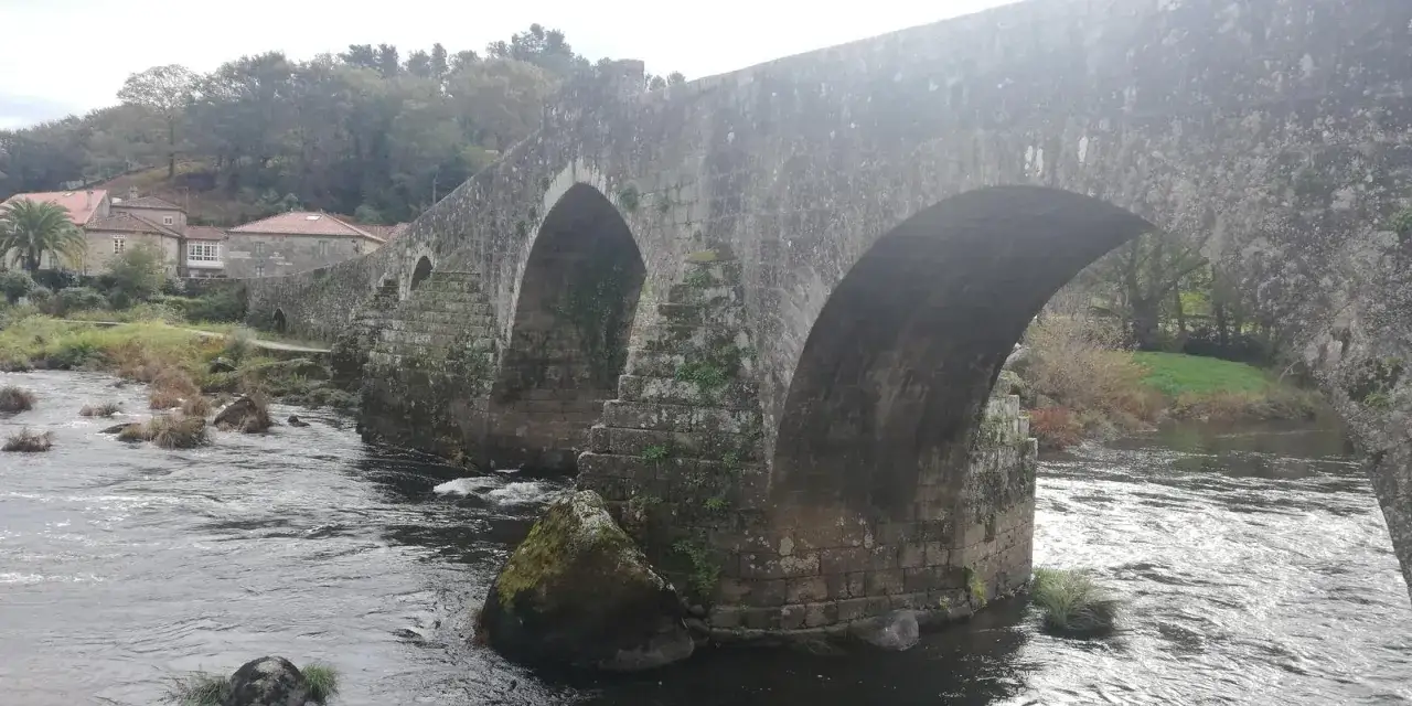 El puente de piedra ponte maceira cruza un río con vegetación y casas al fondo.