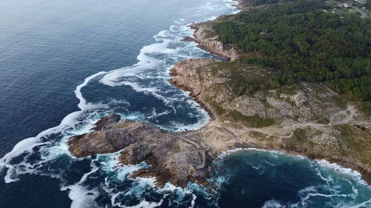 Vista aérea del castro de Baroña, un asentamiento fortificado en la costa gallega, con el mar agitado y espuma blanca.