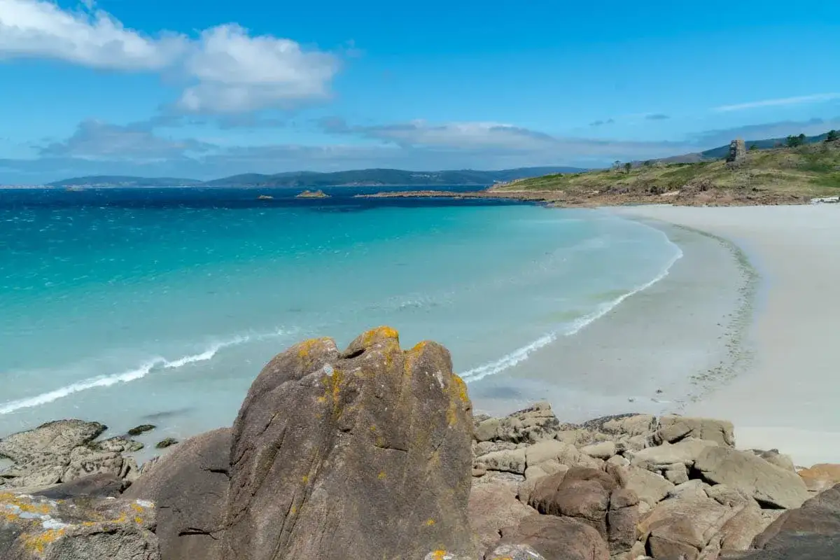 Aguas turquesas y arena blanca en las playas de Finisterre. Rocas cubiertas de líquenes en primer plano, con el mar azul y el cielo nublado al fondo.