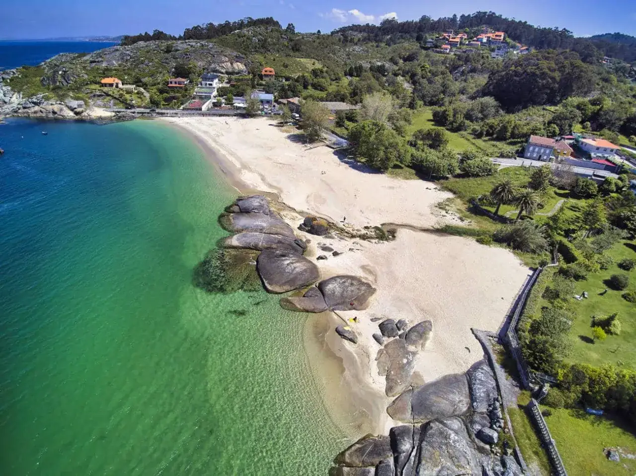 Vista aérea de la playa pintens, con arena dorada, aguas cristalinas y rocas. Casas y vegetación verde cubren las colinas circundantes.