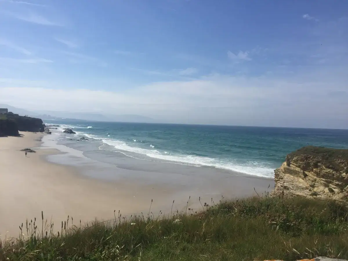 Un paseo solitario por las playas de Lugo, con el mar turquesa rompiendo suavemente en la arena dorada y acantilados verdes.