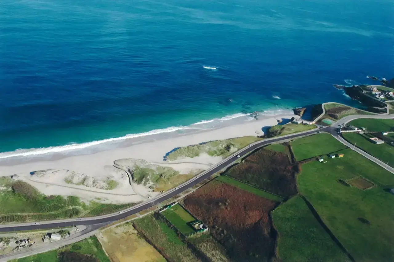 Vista aérea de la playa llas con arena blanca, mar azul y campos verdes. Una carretera serpentea junto a la costa.