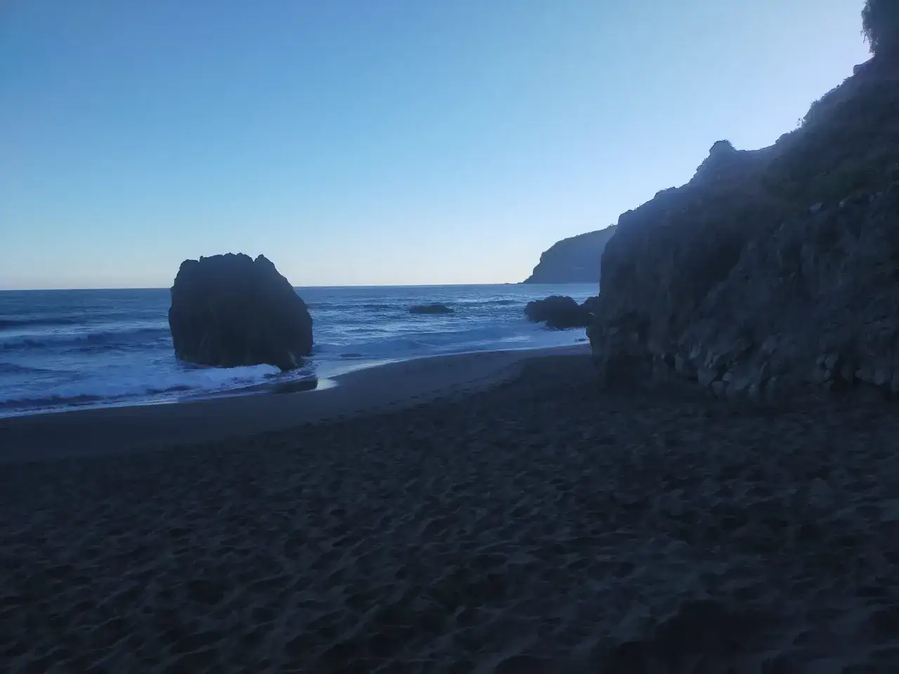 Arena oscura y rocas en la playa los patos al amanecer. El mar azul se une a la costa, con huellas solitarias en la arena.