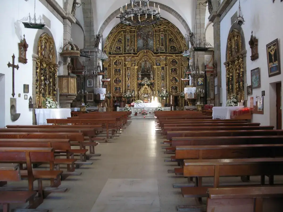 Interior de una iglesia con bancos de madera y un retablo dorado. Una pequeña barca cuelga del techo, evocando la Virxe da Barca.