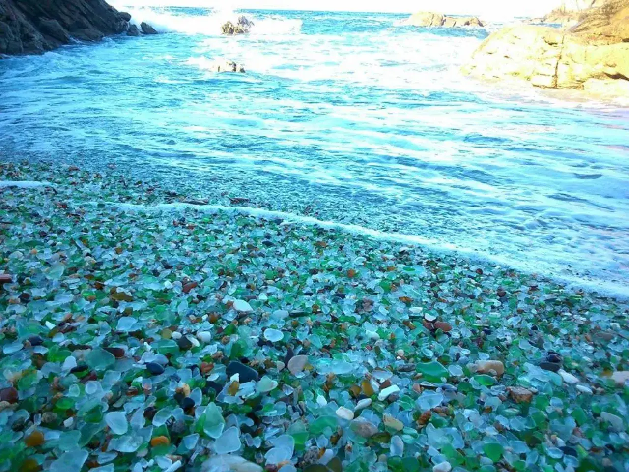 La praia dos cristais brilla con miles de fragmentos de vidrio pulido por el mar, mezclados con guijarros, mientras las olas acarician la costa.