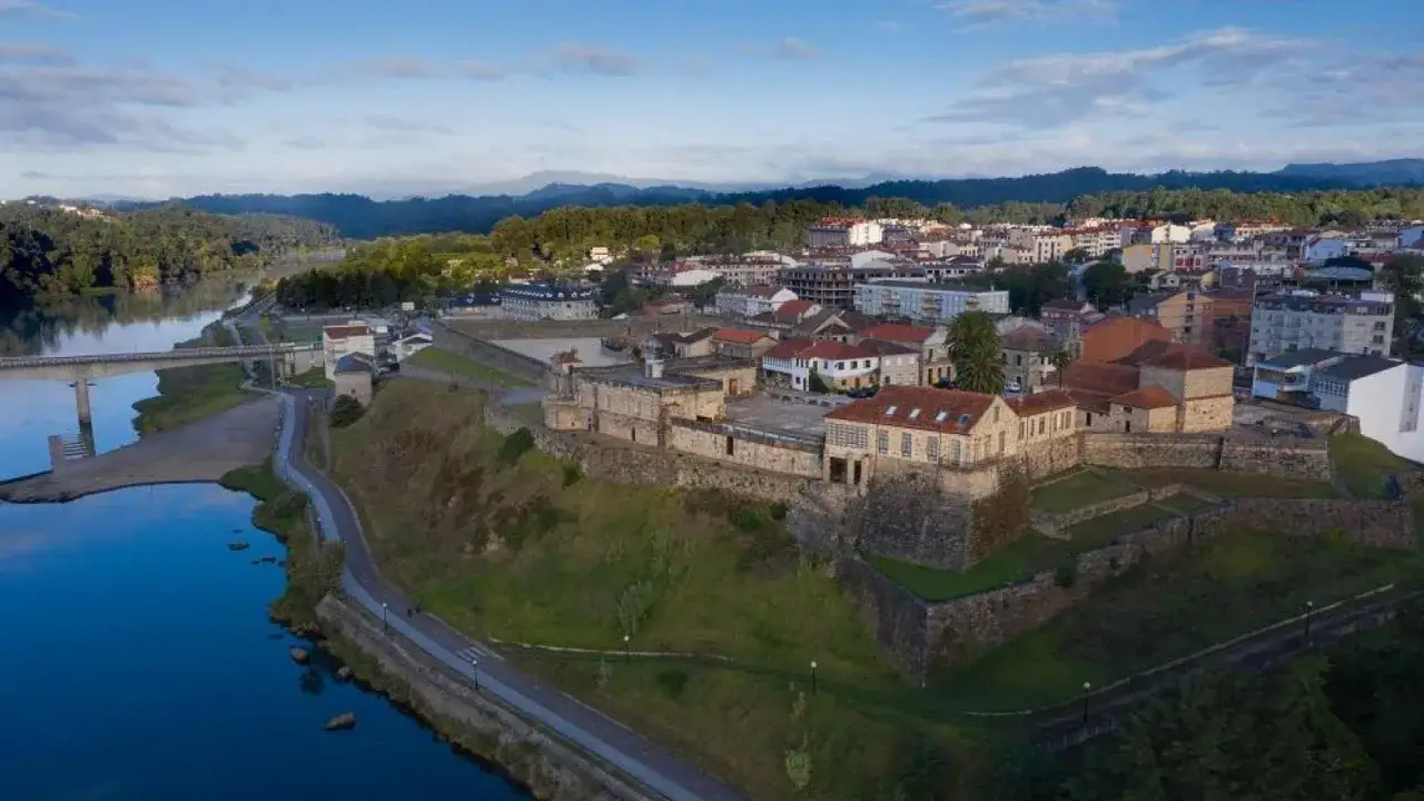 Vista aérea de la torre de Lapela, una fortaleza histórica junto a un río, con un pueblo al fondo y montañas cubiertas de vegetación.