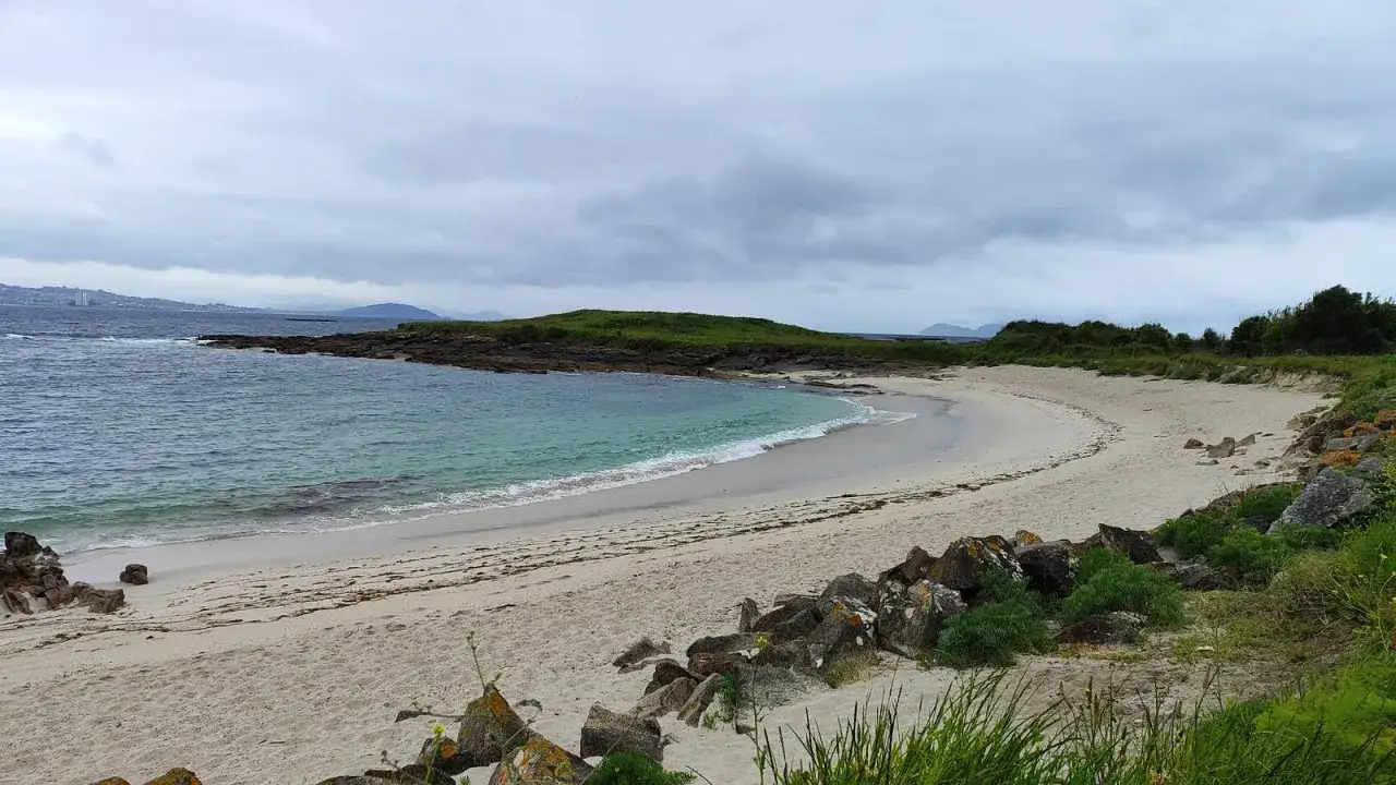 La serena playa Santa Marta de Cangas, con arena dorada y aguas turquesas, se extiende bajo un cielo nublado.