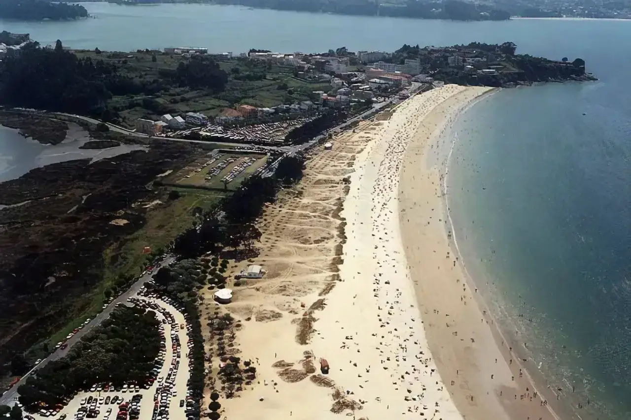 Vista aérea de una playa grande y concurrida, con arena dorada, mar azul y un pueblo costero al fondo.