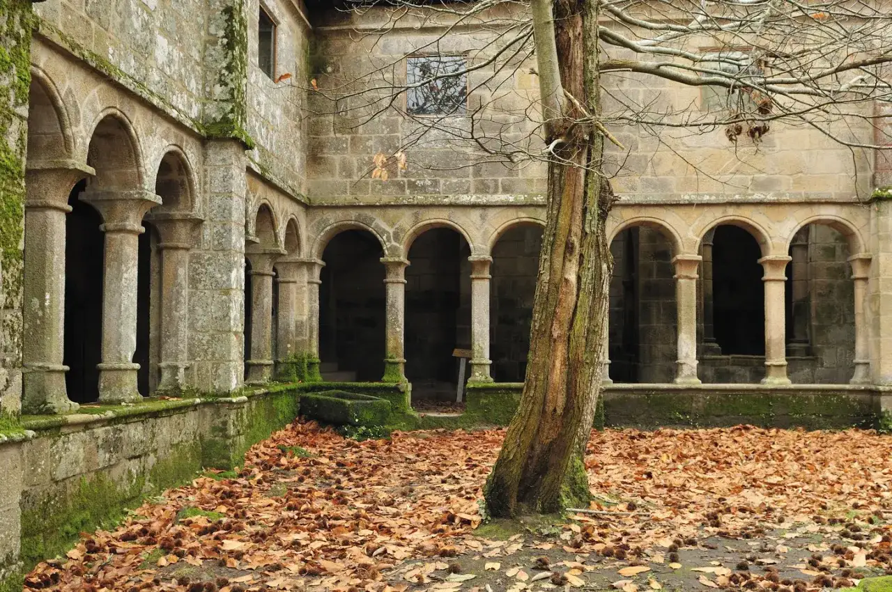 Claustro de piedra cubierto de musgo, con arcos y columnas, rodeado de hojas secas. Un árbol solitario se alza en primer plano, evocando la atmósfera de los monasterios de Galicia.