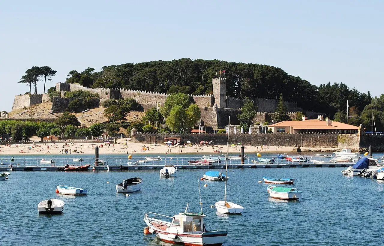 La fortaleza de Monterreal se alza majestuosa sobre una colina, vigilando un puerto lleno de barcas y una playa con gente disfrutando del sol.