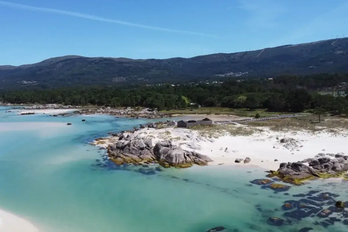 Aguas turquesas bañan la playa Caolín, con arena blanca, rocas y pinares al fondo.