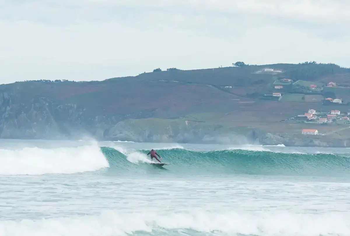 Surfista desliza sobre una ola en la playa de Frouxeira, con colinas verdes y casas al fondo.