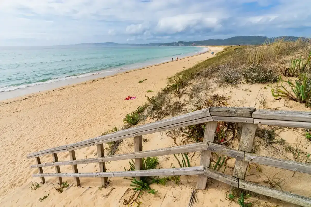 Escalera de madera hacia la playa a lanzada, con arena dorada, mar turquesa y dunas cubiertas de vegetación.