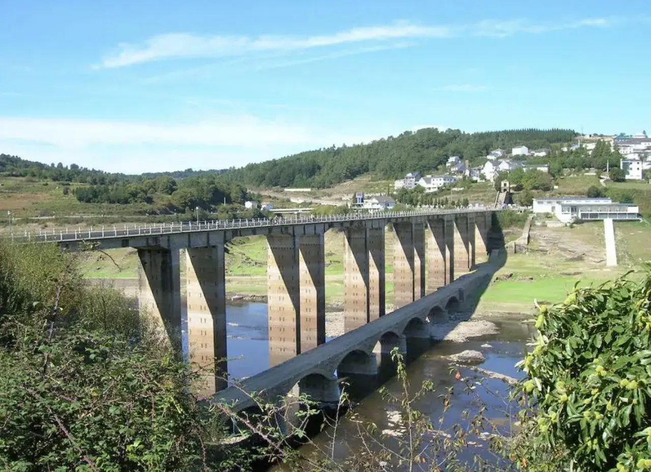 El puente Portomarín cruza un río con un segundo puente más antiguo debajo. El paisaje es verde y montañoso, con casas blancas en la ladera.